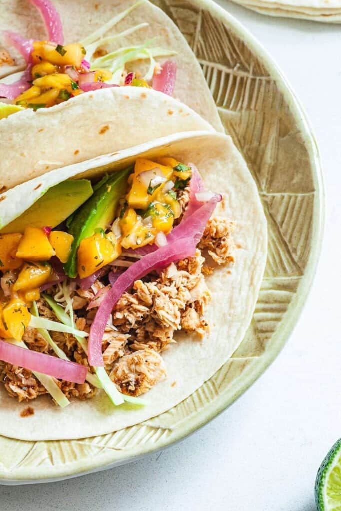Canned salmon tacos served in soft tortillas filled with flaked salmon, shredded cabbage, avocado slices, pickled red onions, and fresh mango salsa. The tacos sit on a patterned plate with bright toppings adding color and texture to the salmon filling.
