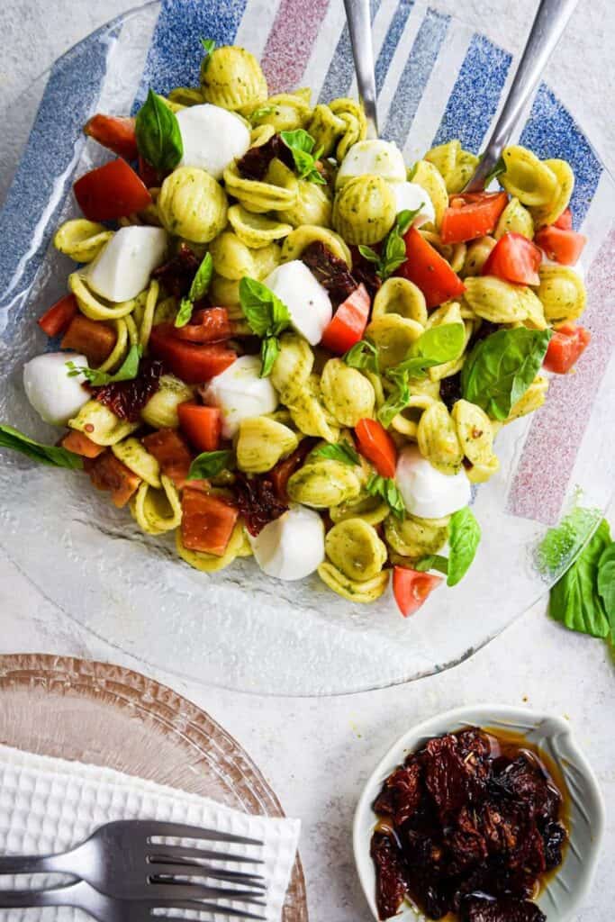 Fresh Caprese pasta salad served on a clear glass platter, featuring orecchiette pasta tossed in pesto with cherry tomatoes, sun-dried tomatoes, mini mozzarella balls, and fresh basil leaves. The vibrant salad is accompanied by a small dish of sun-dried tomatoes in oil and a plate with forks and a napkin nearby, set on a light surface.