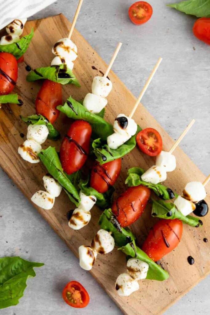 Skewers of Caprese salad arranged on a wooden board, featuring cherry tomatoes, fresh basil leaves, and mozzarella balls drizzled with balsamic glaze. A few halved tomatoes and basil leaves are scattered on the gray surface for a rustic touch.