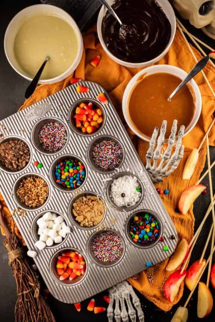 Caramel apple bar with toppings in a muffin tin and bowls of caramel sauce for dipping. 