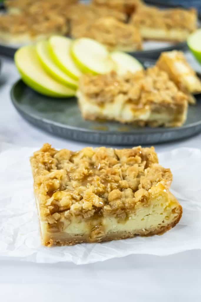 A close-up of a caramel apple cheesecake bar, featuring a crumbly oat topping, creamy cheesecake center, and gooey caramel drizzle. In the background, more bars and fresh green apple slices are served on a dark plate.