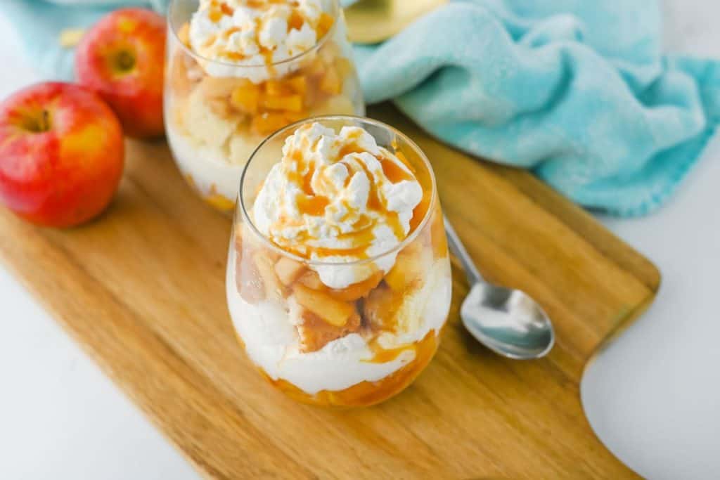 Two glass cups filled with layered caramel apple trifle sit on a wooden board, featuring chunks of spiced apples, whipped cream, cake, and a drizzle of caramel sauce. Fresh apples and a spoon rest nearby, evoking a cozy fall dessert vibe.