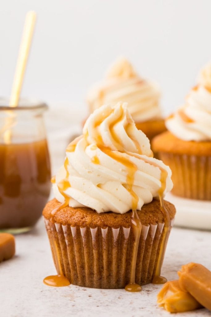 Close-up of a caramel cupcake topped with a swirl of vanilla buttercream and drizzled with rich caramel sauce. The cupcake is surrounded by soft caramel candies and a jar of caramel in the background, with more frosted cupcakes slightly out of focus.