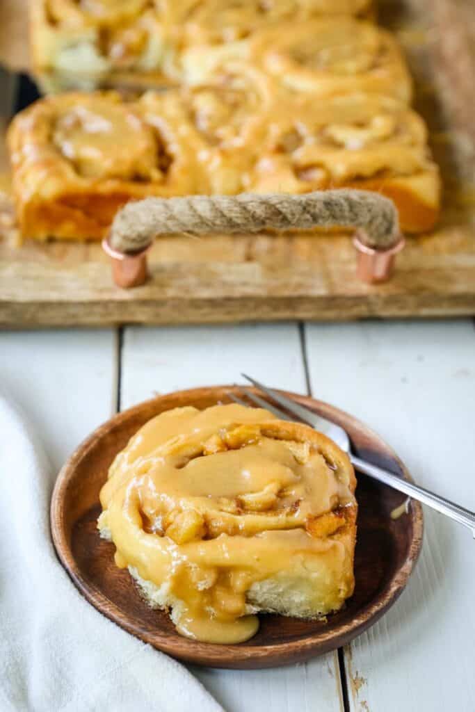 A gooey apple cinnamon roll sits on a wooden plate, covered in thick caramel glaze with tender apple pieces peeking through the soft, swirled dough. In the background, a tray of fresh rolls rests on a rustic wooden serving board.