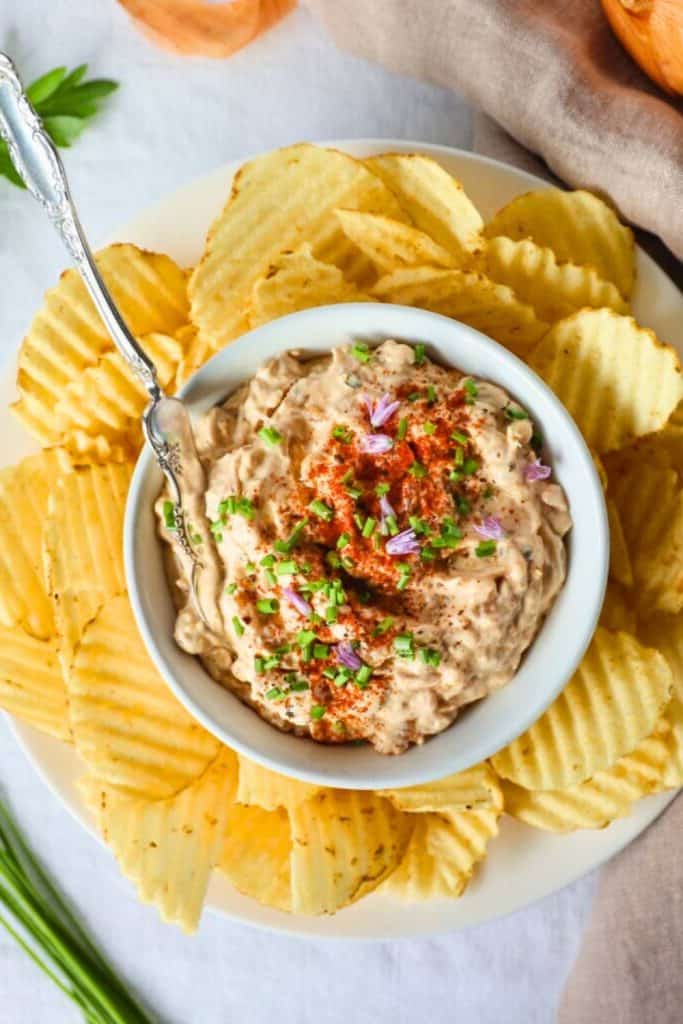 Caramelized onion and blue cheese dip served in a small white bowl, topped with chopped chives, paprika, and edible purple flowers. Surrounded by ridged potato chips on a white plate, perfect for a savory game day snack.