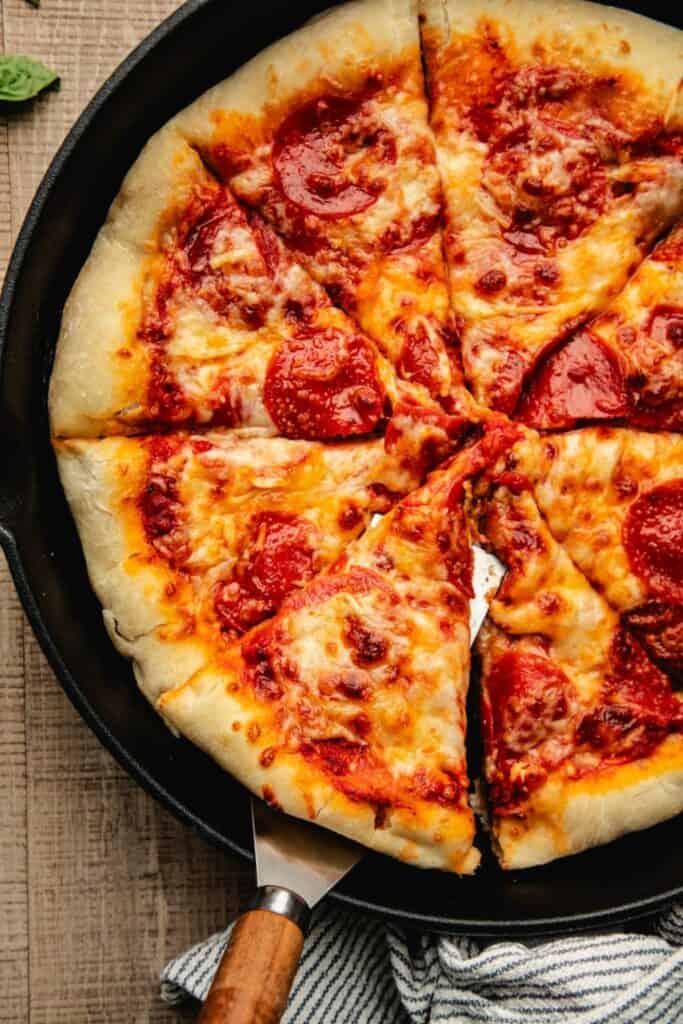 Cast iron skillet pizza topped with melted mozzarella and pepperoni sliced into wedges inside a black skillet. One slice is lifted slightly with a metal spatula showing the thick crust and bubbly cheese.