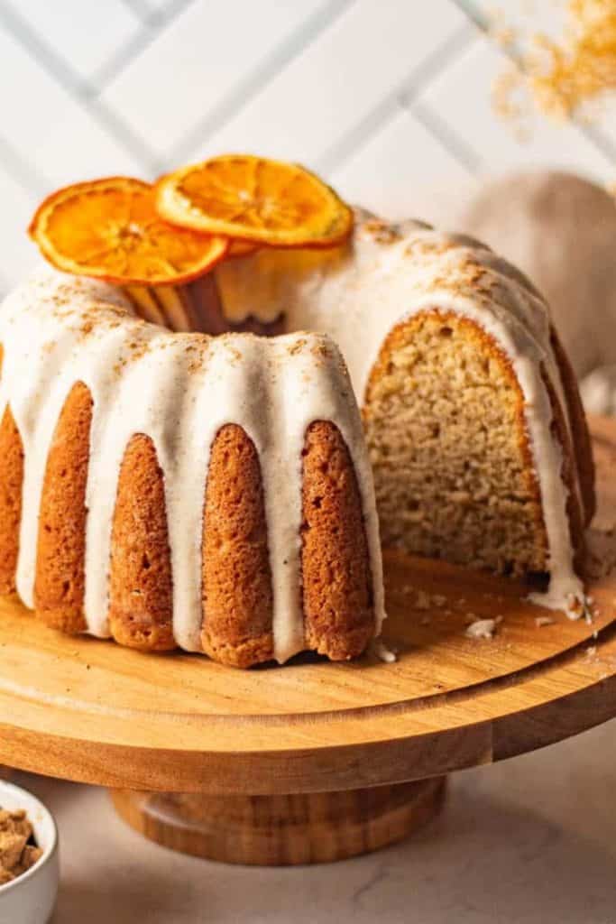 A moist bundt cake with white icing drizzled over the ridges, topped with thin dried orange slices. One slice is removed to reveal the soft, dense crumb inside, and the cake sits on a wooden cake stand against a tiled background.