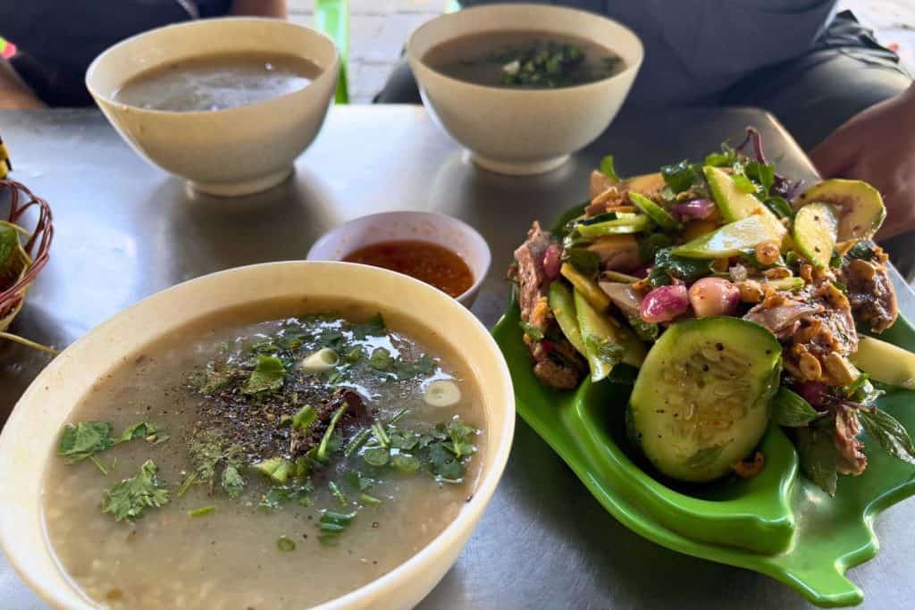 Three bowls of steaming rice porridge garnished with scallions, cilantro, and black pepper sit on a metal table, accompanied by a dipping sauce. Next to them is a vibrant plate of Vietnamese duck salad with sliced cucumbers, herbs, onions, and roasted peanuts, served on a bright green dish.