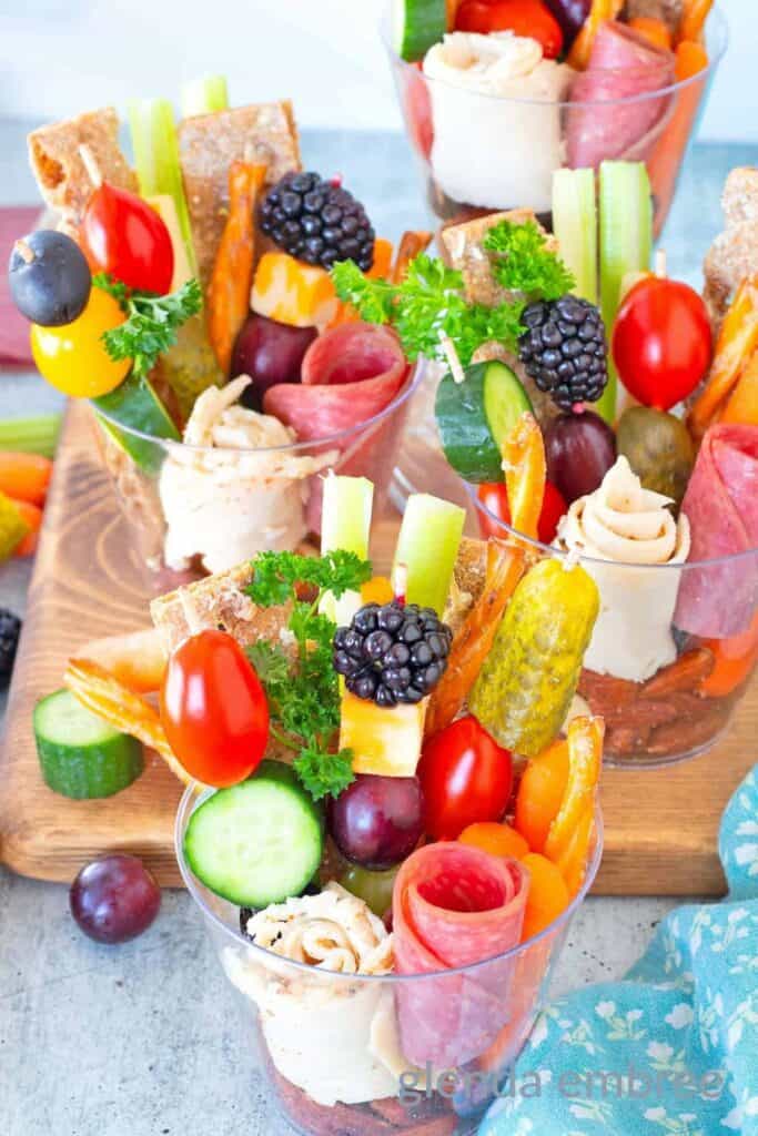 Clear cups filled with charcuterie including rolled deli meats, cheese cubes, crackers cucumber slices, grapes cherry tomatoes, olives and pickles arranged vertically for easy snacking. The colorful layered ingredients make these quick road trip snacks for teens