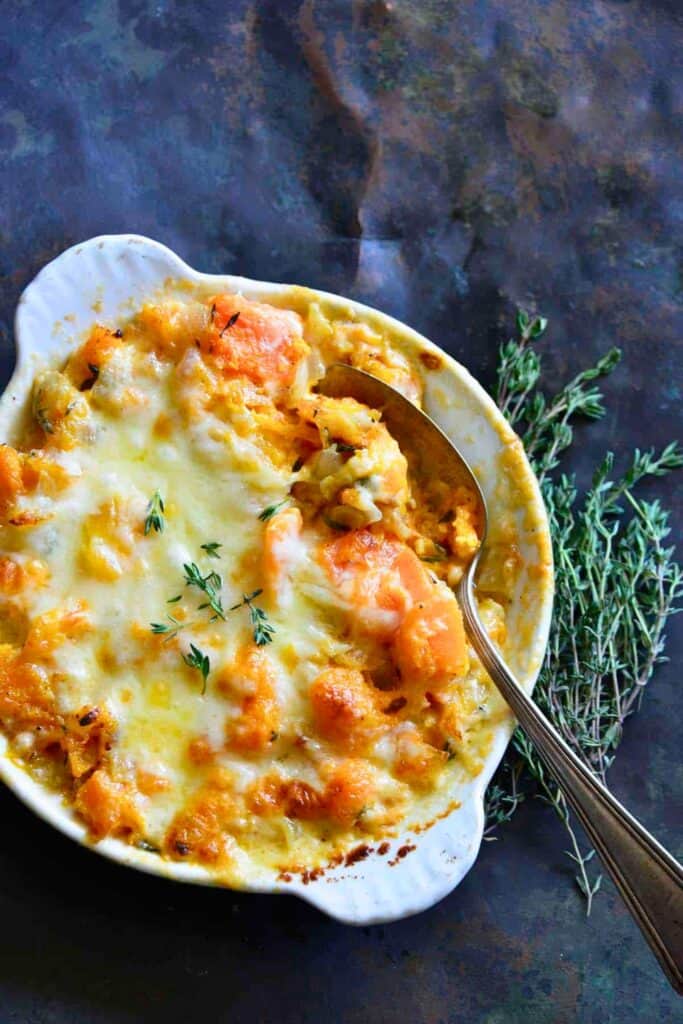 Cheesy butternut squash gratin with tender squash pieces, melted golden cheese and fresh thyme in a white baking dish, a cozy option for make ahead fall casserole ideas.