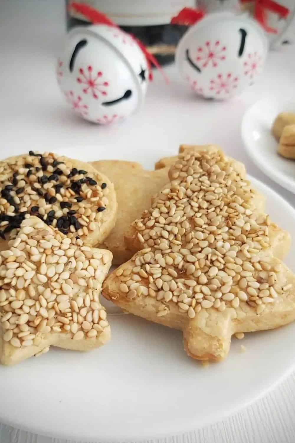 Turkish cheese biscuits shaped like stars and rounds and generously coated with white and black sesame seeds. The golden savory biscuits are arranged on a white plate with decorative bells in the background.