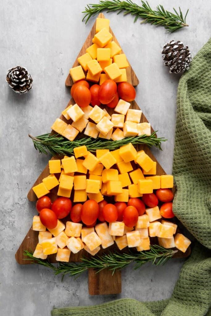 Cubed cheese board on a Christmas Tree shaped wooden cutting board with rows of alternating cheese cubes, cherry tomatoes and rosemary sprigs.