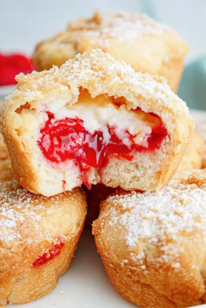 Alt text: Cherry cheesecake muffins with a soft golden exterior filled with creamy cheesecake and bright red cherry filling dusted with powdered sugar. One muffin is split open to reveal the rich center while others are stacked underneath.