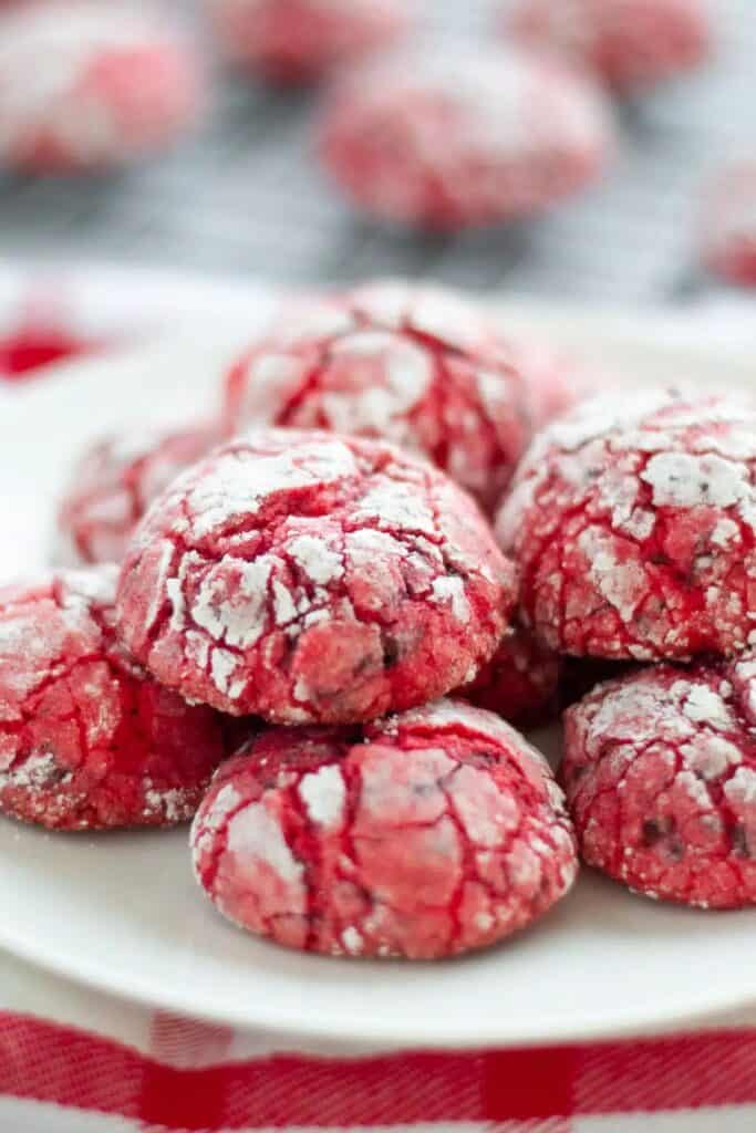 Close-up of red chocolate crinkle cookies with cherries, dusted generously in powdered sugar to highlight the cracked texture. The cookies are stacked on a white plate with more cooling in the blurred background, adding a festive holiday vibe.