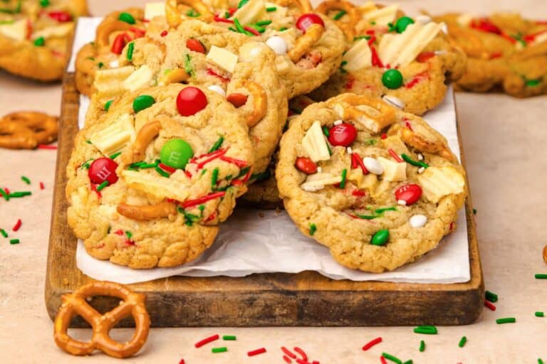 Chewy Kitchen Sink Christmas cookies stacked on a wooden board, loaded with red and green M&Ms, white chocolate chunks, pretzel twists, potato chips, and holiday sprinkles, with scattered sprinkles and pretzels on the table.