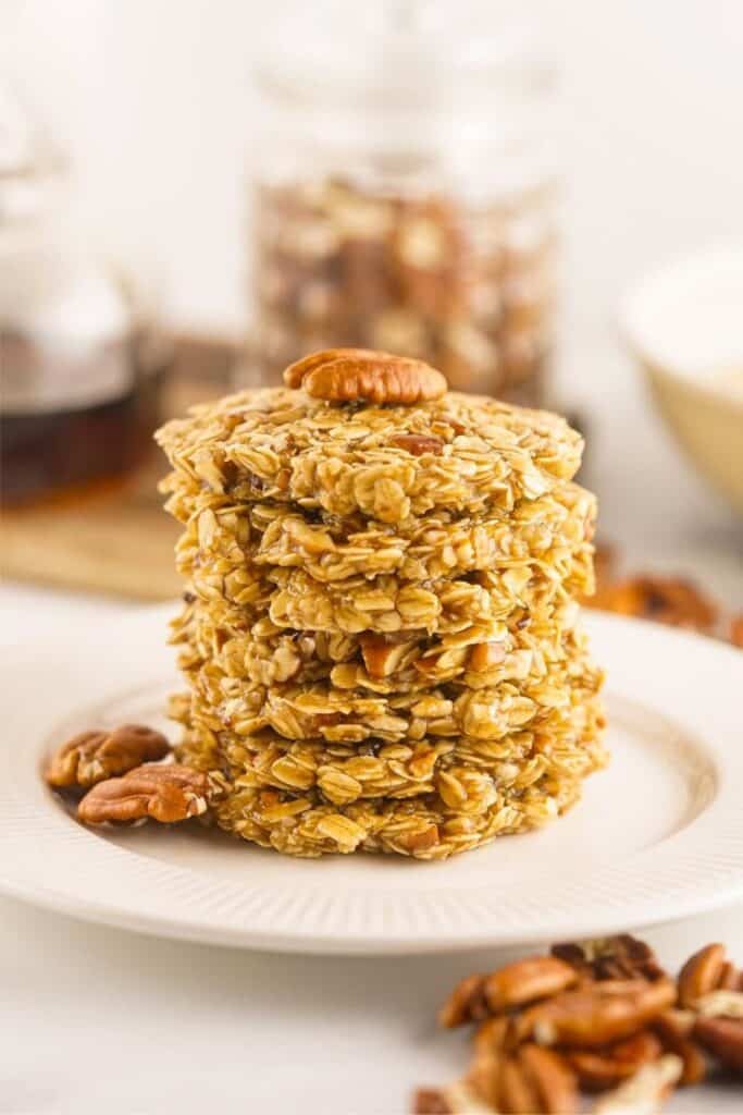 A tall stack of golden Maple Pecan No Bake Cookies made with visible rolled oats and chopped pecans sits on a white plate. A whole pecan tops the stack, with additional pecans scattered on the plate and table around it.