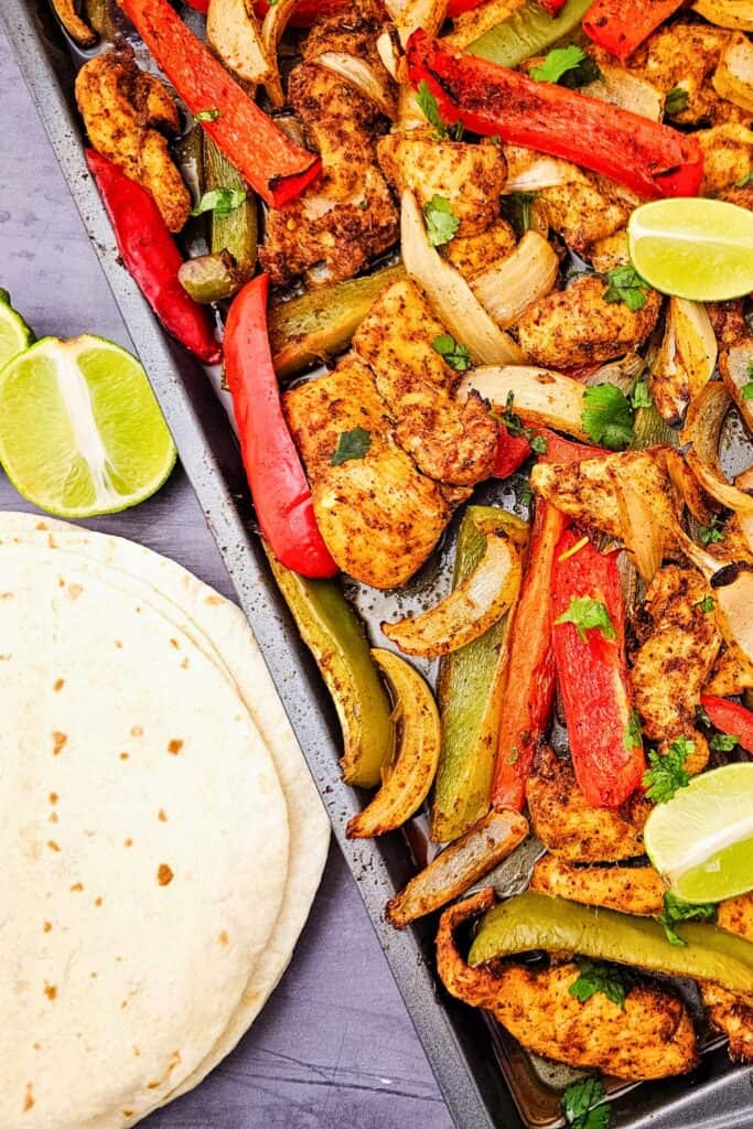 Sheet pan chicken and bell pepper recipe featuring seasoned roasted chicken strips with red and green bell peppers and sliced onions. The pan is garnished with fresh cilantro and lime wedges and served with soft tortillas for quick dinner.