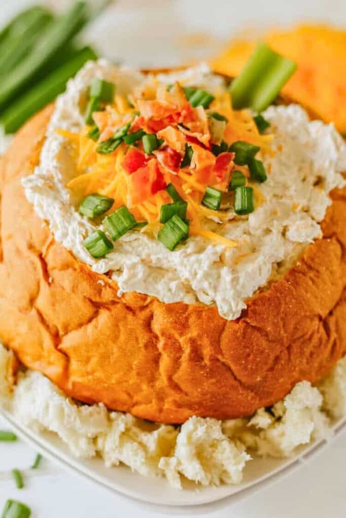 Creamy Chicken Bacon Ranch Crack Dip served in a hollowed-out round bread bowl, topped with shredded cheddar cheese, crispy bacon bits, and chopped green onions. Surrounded by chunks of bread for dipping.