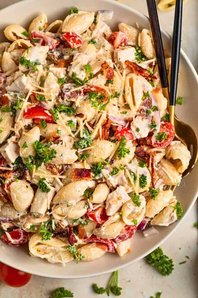 Creamy chicken bacon ranch pasta salad served in a white bowl, featuring shell pasta tossed with crispy bacon, shredded cheese, cherry tomatoes, red onion, chunks of chicken, and fresh parsley. The salad is coated in a rich ranch dressing, making it a hearty and flavorful dish perfect for gatherings.