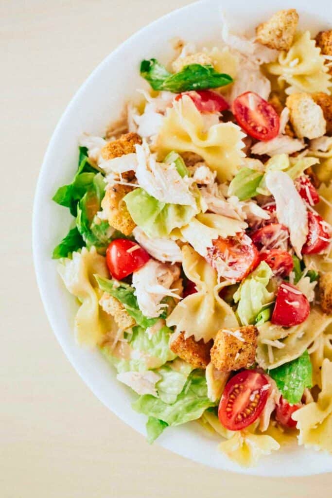 Bowl of chicken Caesar pasta salad with bow tie pasta grilled chicken romaine lettuce cherry tomatoes croutons and shaved parmesan tossed in creamy Caesar dressing.
