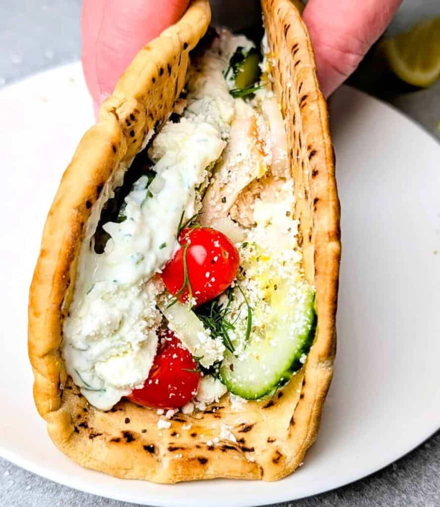 Chicken gyros served in warm grilled pita bread filled with sliced chicken, cucumber, cherry tomatoes, fresh herbs, and creamy tzatziki sauce. The pita wrap is held open to show the colorful fillings and crumbled cheese inside.