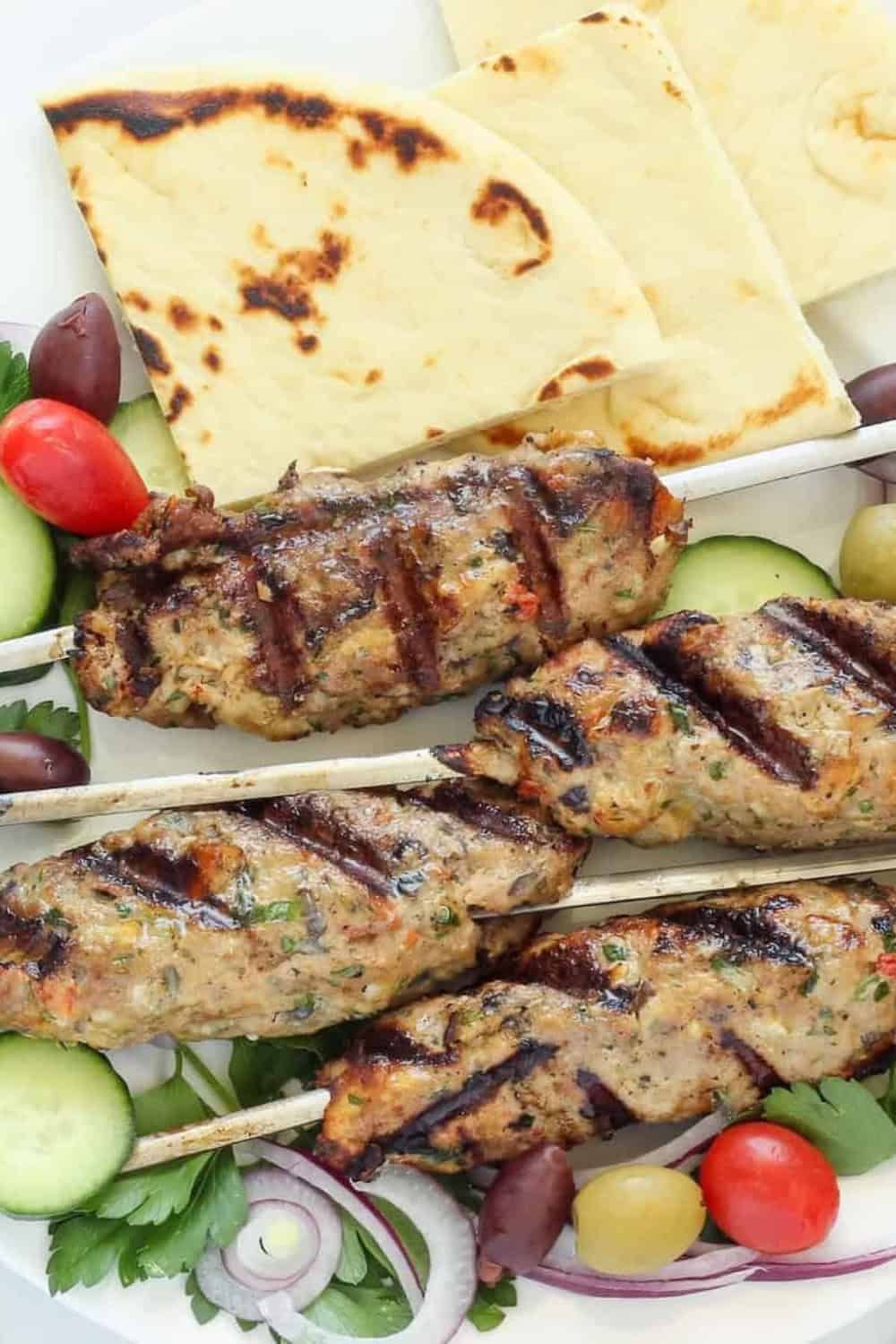 Grilled chicken kofta skewers with char marks arranged on a platter with cucumber slices, cherry tomatoes, red onion, olives, and fresh parsley. Warm pita bread is served alongside the skewers, creating a colorful summer meal.