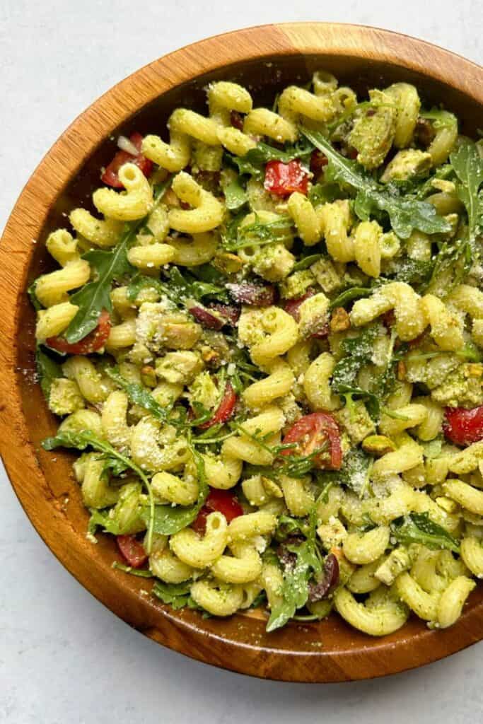 Chicken pesto pasta salad served in a wooden bowl, featuring cavatappi pasta tossed with chunks of chicken, cherry tomatoes, arugula, Kalamata olives, pistachios, and a generous coating of green pesto. The dish is finished with a sprinkle of grated Parmesan cheese for a flavorful and vibrant presentation.