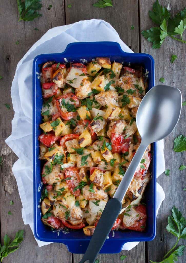 Chicken potato casserole bake with chunks of seasoned chicken, roasted potatoes, red bell peppers, melted cheese and fresh parsley in a blue rectangular baking dish.