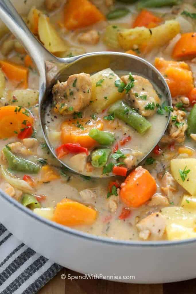 A metal serving spoon full of chicken stew is held over a hearty bowl of the stew with chuncks of chicken, peas, potatoes and carrots visible.