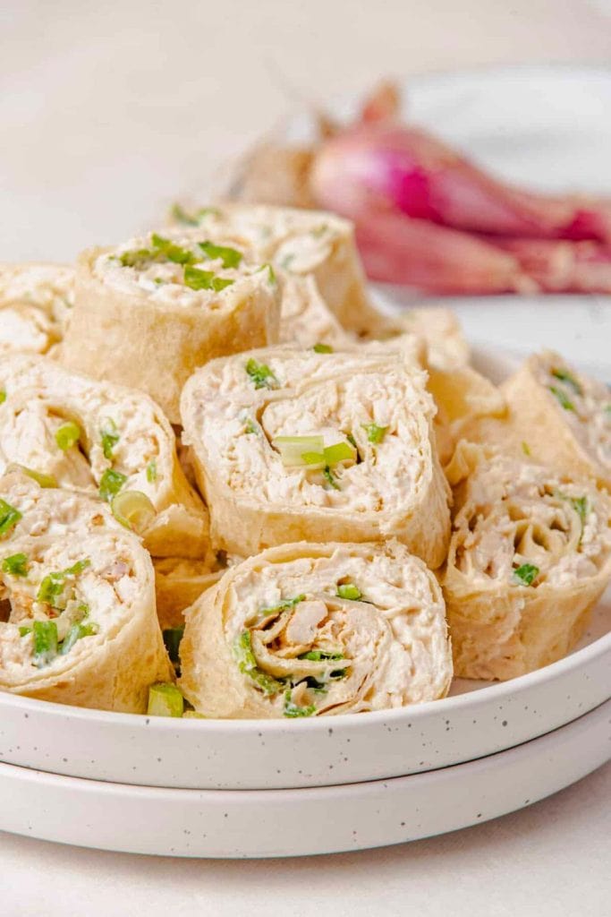 Close-up of tortilla pinwheels filled with a creamy chicken and green onion mixture, sliced and stacked on a white speckled plate. This easy tailgate food is served cold and rolled tightly for convenient bite-sized portions.