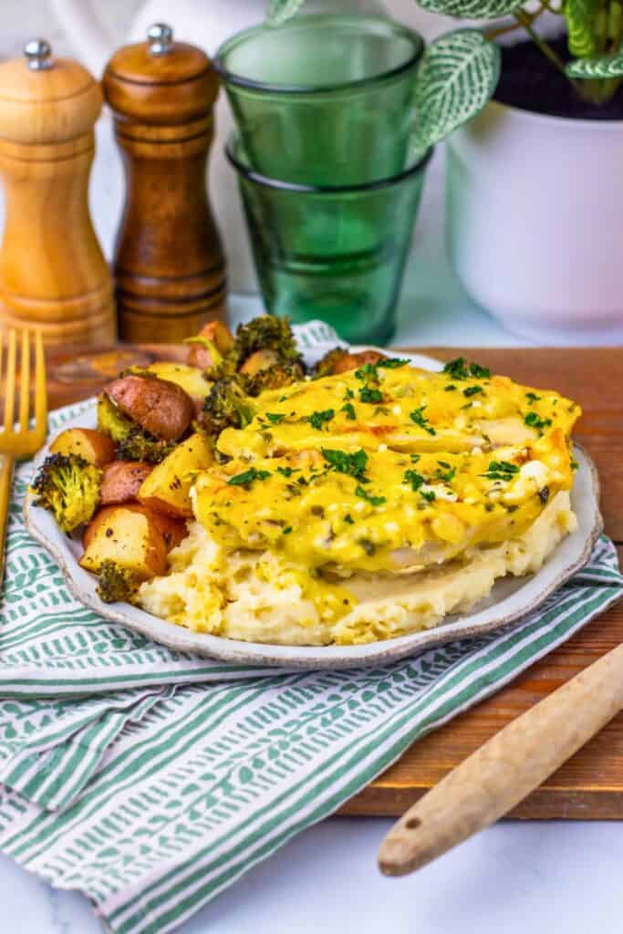 Chicken and vegetables sheet pan dinner served on mashed potatoes, topped with melted cheese and parsley, with a side of roasted red potatoes and broccoli. The plate sits on a green and white striped napkin next to a wooden knife and gold fork.