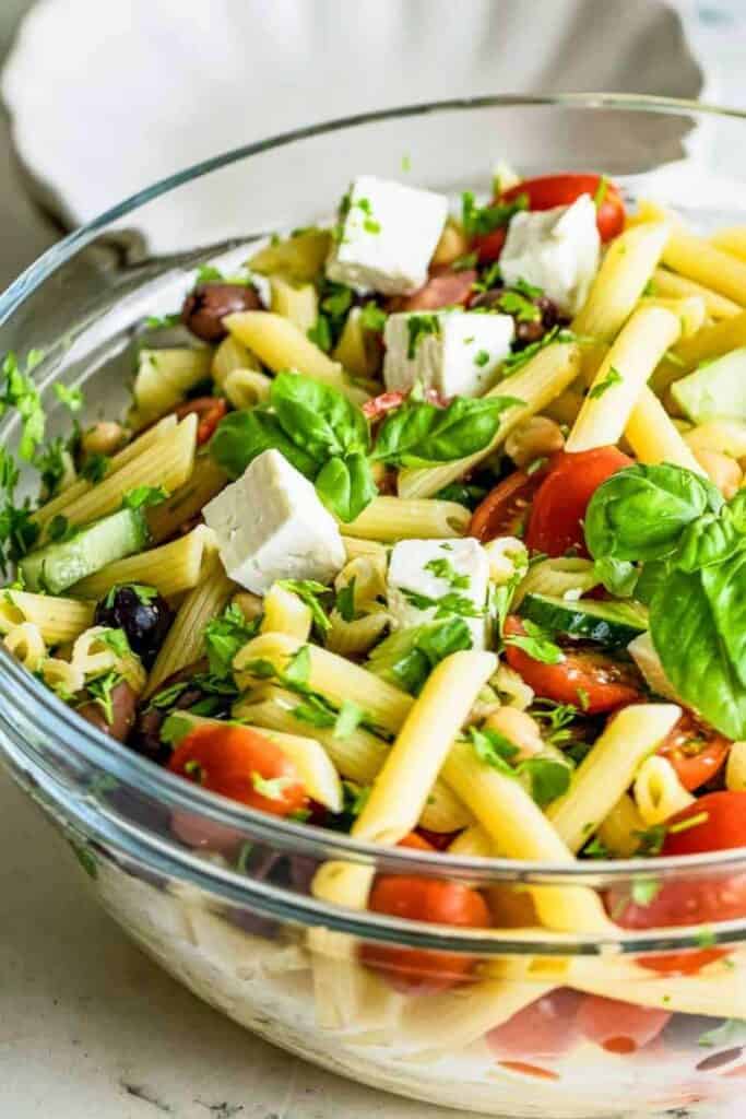 A glass bowl filled with a healthy cold pasta salad made with penne pasta, cherry tomatoes, cucumber, chickpeas, kalamata olives, fresh basil leaves, and cubes of feta cheese, all tossed in a light vinaigrette and garnished with chopped parsley.