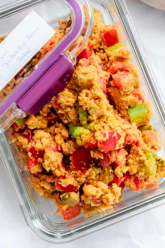 Glass meal prep container filled with mashed chickpea salad mixed with diced red bell pepper, celery, and green onions. The chunky, creamy mixture makes a colorful and protein-packed plant-based option perfect for sandwiches, wraps, or easy lunches.