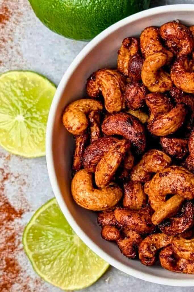 Bowl of chili lime cashews coated in a deep reddish brown seasoning with fresh lime halves arranged beside the bowl. The close up view highlights the crunchy roasted nuts and bright citrus flavors that make this a healthy road trip snack.