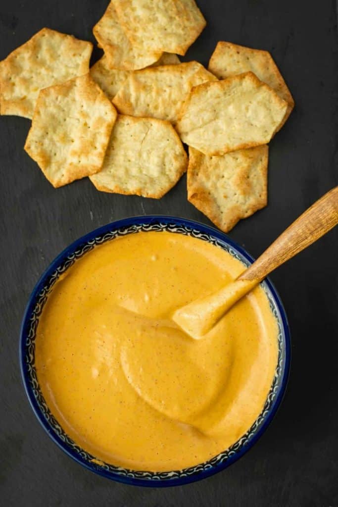 Smooth and creamy chipotle almond dip in a decorative blue bowl, stirred with a wooden spoon. Served with crispy hexagon-shaped crackers on the side.