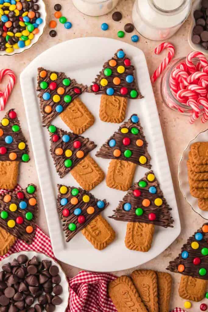 Christmas tree-shaped cookies made from Biscoff biscuits as trunks and piped chocolate topped with colorful M&M candies as tree branches. The cookies are arranged on a white platter surrounded by festive candy canes, chocolate chips, and red gingham cloth.