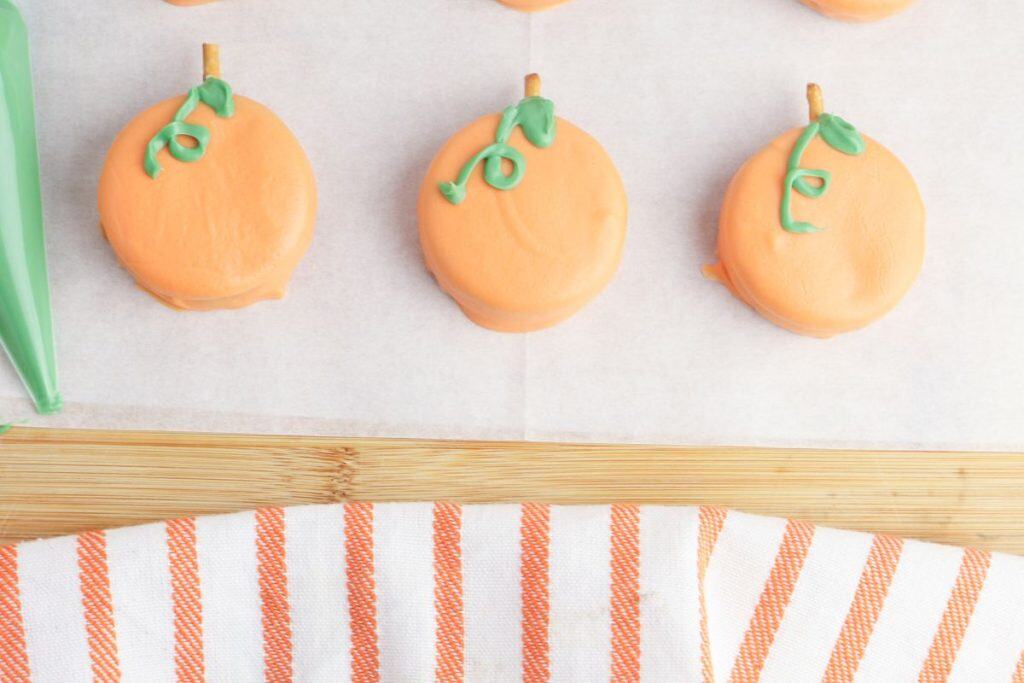 Three DIY pumpkin treats for Halloween made from orange candy-coated oreo cookies, each topped with a pretzel stick stem and green icing vine. The treats are arranged on parchment paper beside a green icing piping bag, with a striped orange and white kitchen towel below.