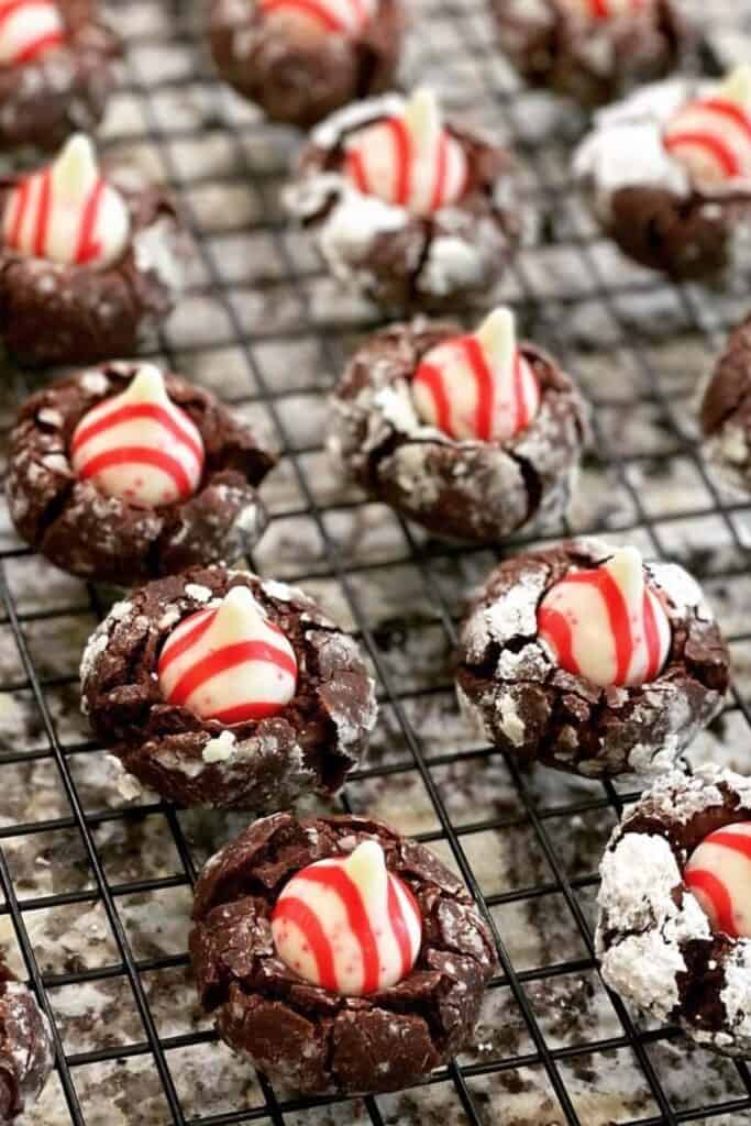 Chocolate Christmas cookies with a crackled powdered sugar coating are topped with red and white striped peppermint Hershey’s Kisses and arranged on a cooling rack.