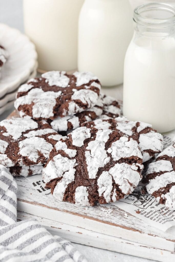 Stack of classic chocolate crinkle cookies coated in powdered sugar, showing their signature cracked surface. The cookies are styled with glass bottles of milk and a striped napkin beside them.