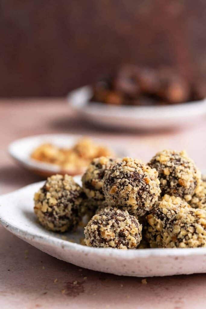 Plate of chocolate date balls rolled in finely chopped nuts, with a rich dark chocolate center and a textured nut coating. The no bake chocolate date balls look dense and fudgy, making them a wholesome sweet treat or easy energy snack.
