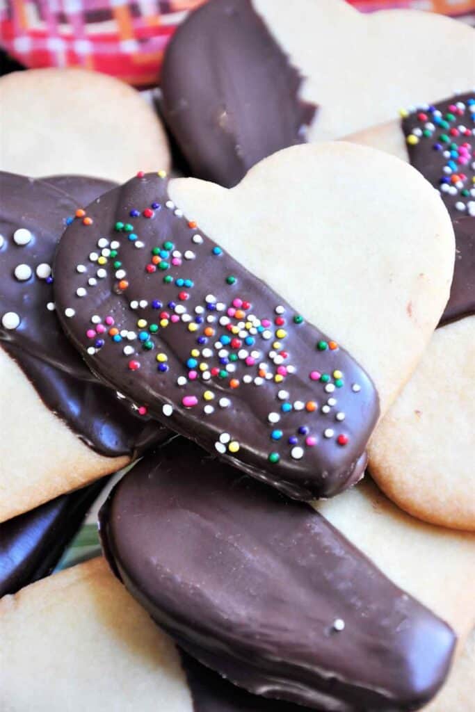 pile of heart shaped vanilla cookies, half dipped with chocolate and rainbow sprinkles. 