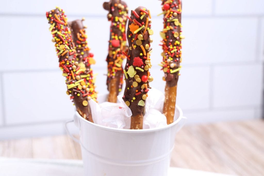 White pail filled with Chocolate Dipped Pretzel Rods, each coated in milk chocolate and covered with festive fall-colored sprinkles in red, orange, yellow, and gold. The rods are displayed upright in tissue paper, creating a playful and seasonal autumn treat.