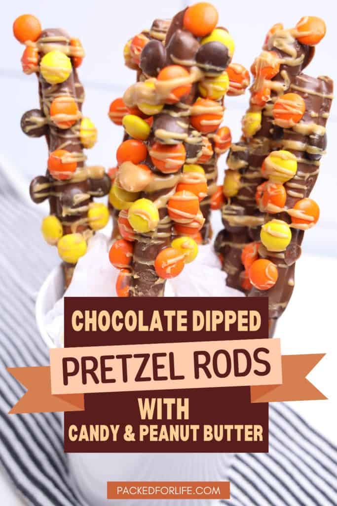 A white cup holds chocolate dipped pretzel rods coated with orange, yellow, and brown candy pieces and drizzled with peanut butter.