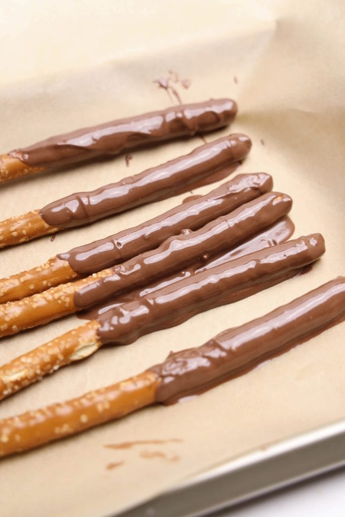 Six chocolate dipped pretzel sticks laid out on parchment paper, with the top half of each pretzel coated in smooth, glossy milk chocolate. The pretzels are arranged in neat rows, ready to set or be decorated.