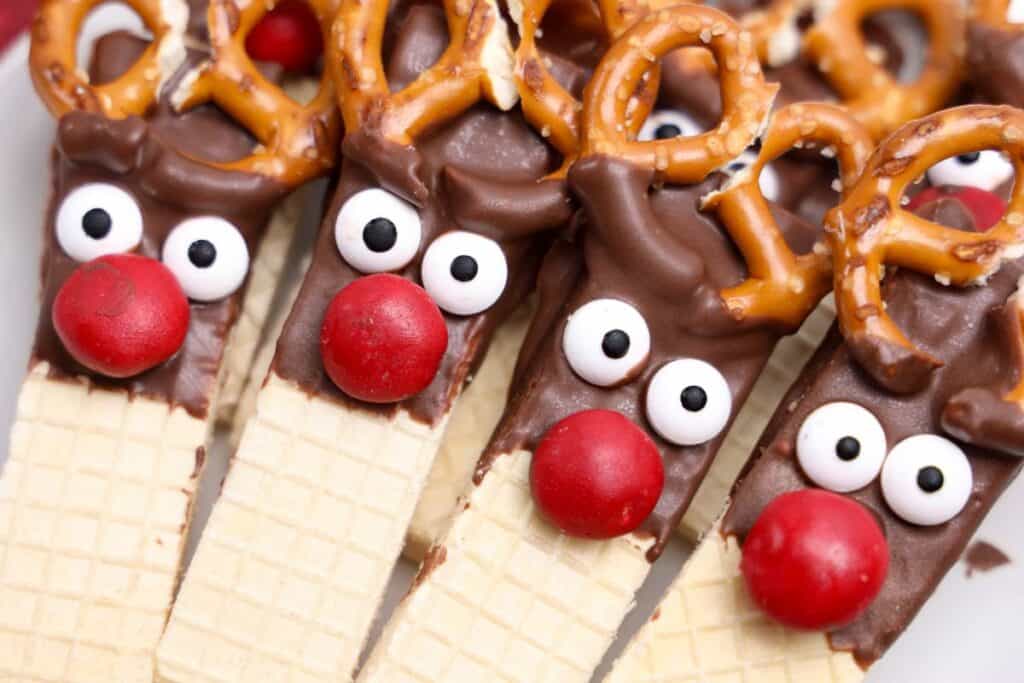 Close-up of chocolate-dipped reindeer cookies made with vanilla wafer bars, featuring candy eyes, red M&M noses, and mini pretzels for antlers. The whimsical holiday treats are tightly arranged, highlighting their detailed and playful decorations.