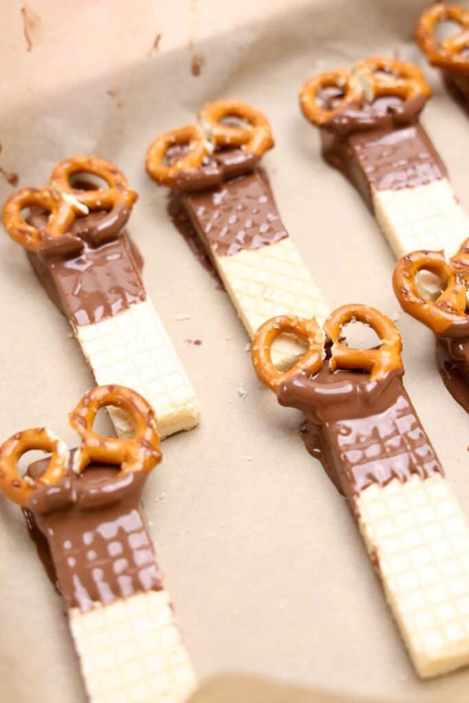 Vanilla wafer cookies partially dipped in melted chocolate and topped with mini pretzels arranged as antlers, laid out on parchment paper to set. These cookies are in the early decorating stage of becoming reindeer treats.
