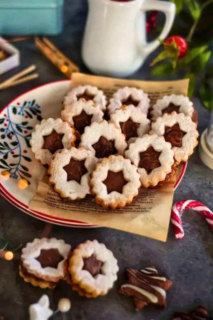 Festive plate of sandwich-style chocolate cookies with scalloped edges and star-shaped cutouts, filled with rich chocolate spread and dusted with powdered sugar. The cookies are styled on parchment paper with candy canes, and holiday decorations.