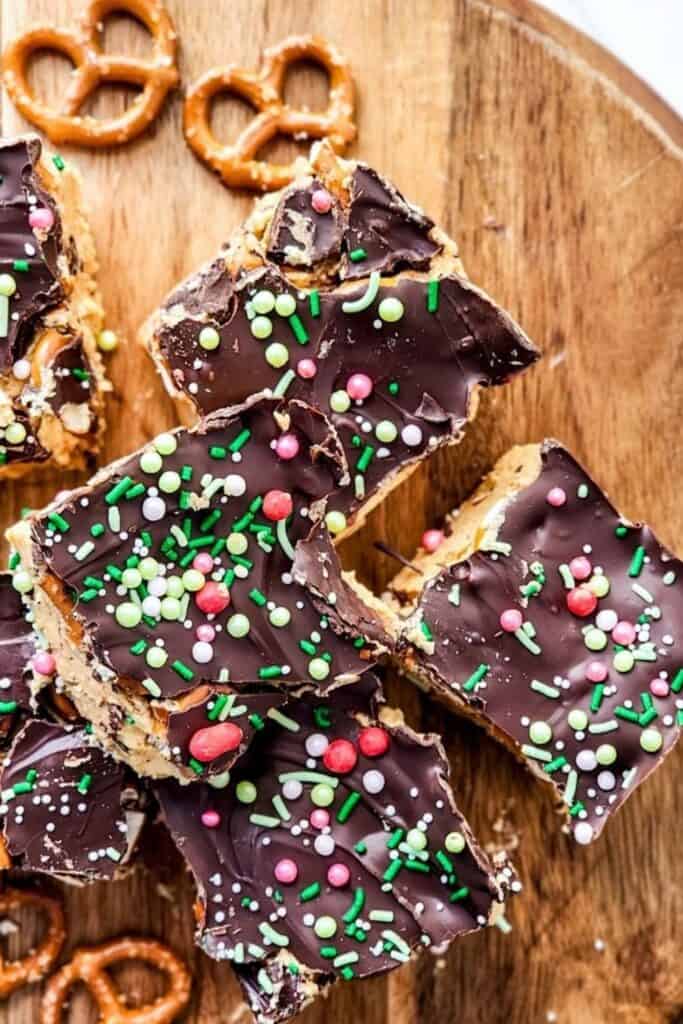 Festive chocolate peanut butter pretzel bars topped with a thick layer of chocolate and colorful holiday sprinkles. The bars are loaded with crunchy pretzels and creamy peanut butter, served on a wooden board for a sweet and salty Christmas dessert.