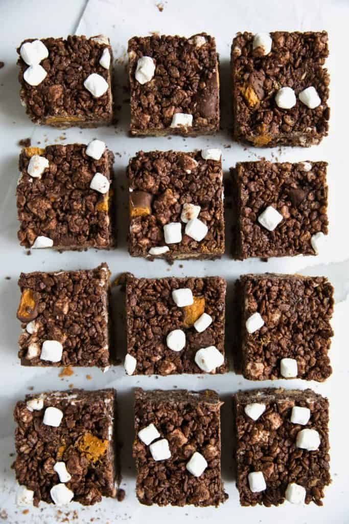 Chocolate peanut butter Rice Krispies treat squares topped with mini marshmallows and chunks of candy bars, arranged in a grid pattern on parchment paper.