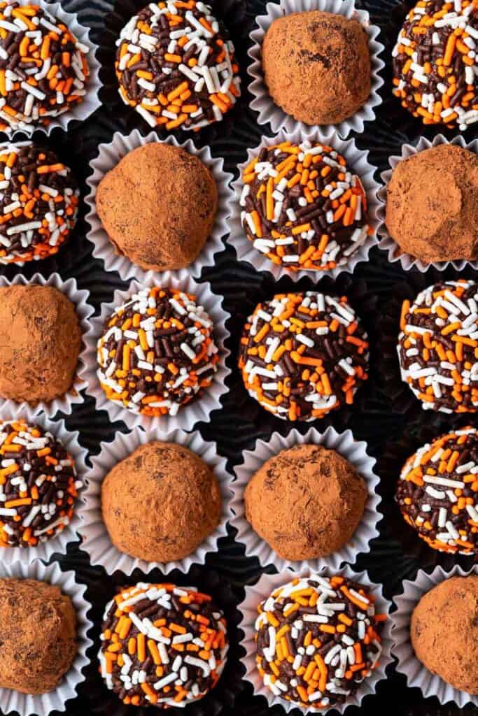 Chocolate pumpkin truffles arranged in individual paper cups with alternating coatings of cocoa powder and orange, white, and chocolate sprinkles.
